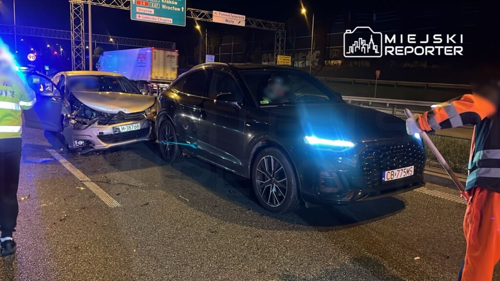 Dwa uszkodzone samochody osobowe stoją na drodze, obok pracownik w odblaskowej kamizelce kieruje ruchem.