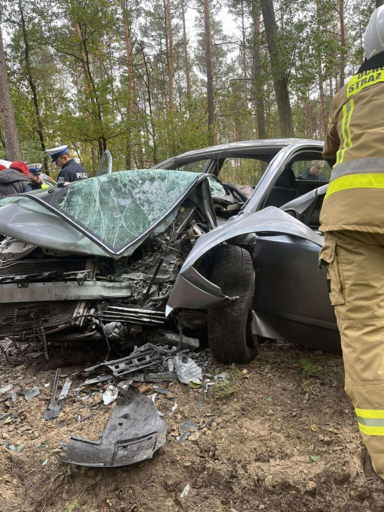 Zniszczony samochód osobowy leży na boku w lesie, obok niego pracują strażacy i policjanci.