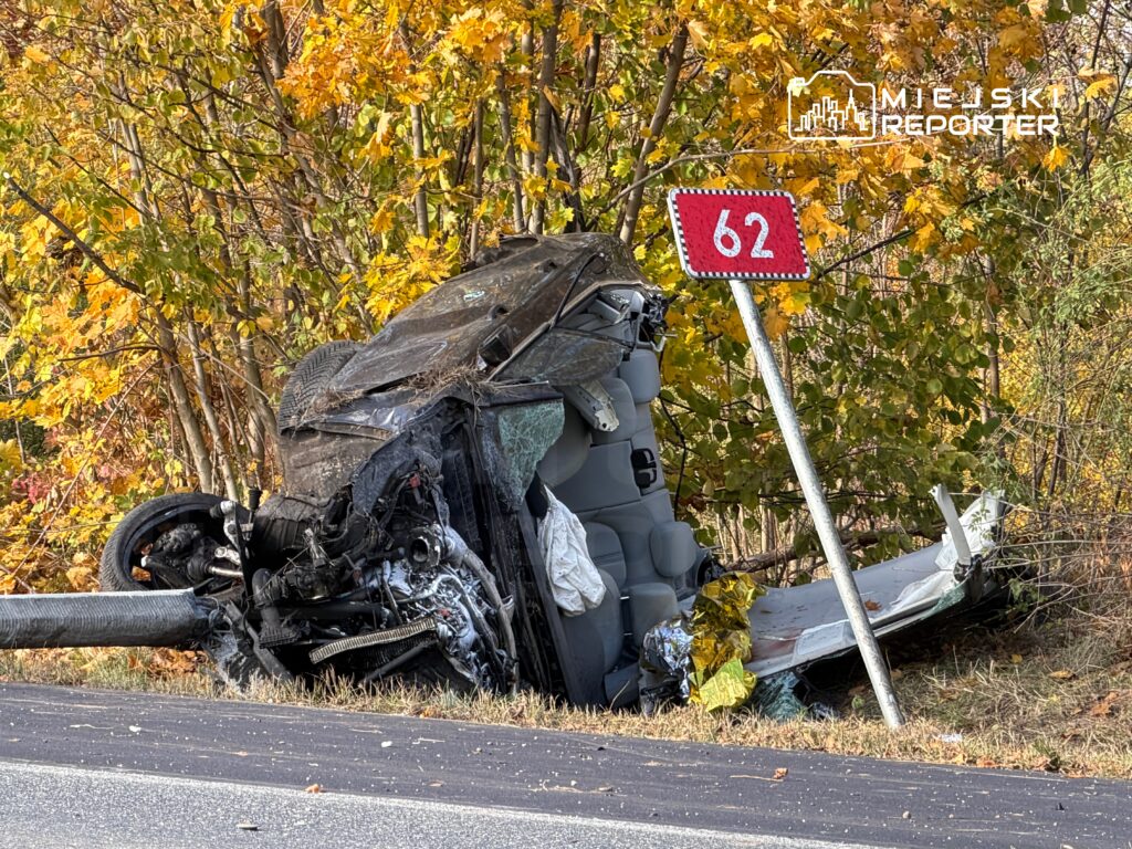 Samochód leży na boku przy drodze, obok znaku drogowym z numerem 62, otoczony kolorowymi liśćmi drzew.