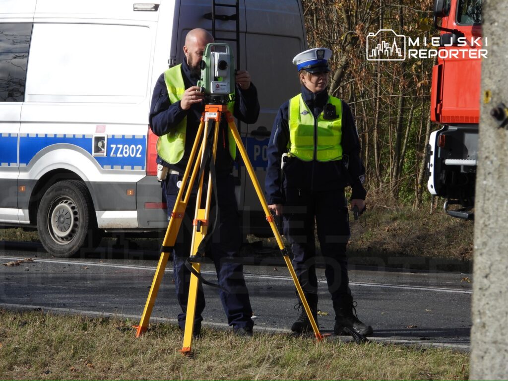 Mężczyzna w kamizelce odblaskowej obsługuje urządzenie pomiarowe, a obok stoi policjantka z radiotelefonem na drodze.