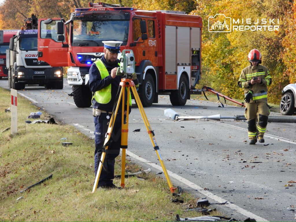 Policjant obsługujący sprzęt pomiarowy na drodze, w tle pojazdy straży pożarnej i reszta służb ratunkowych.