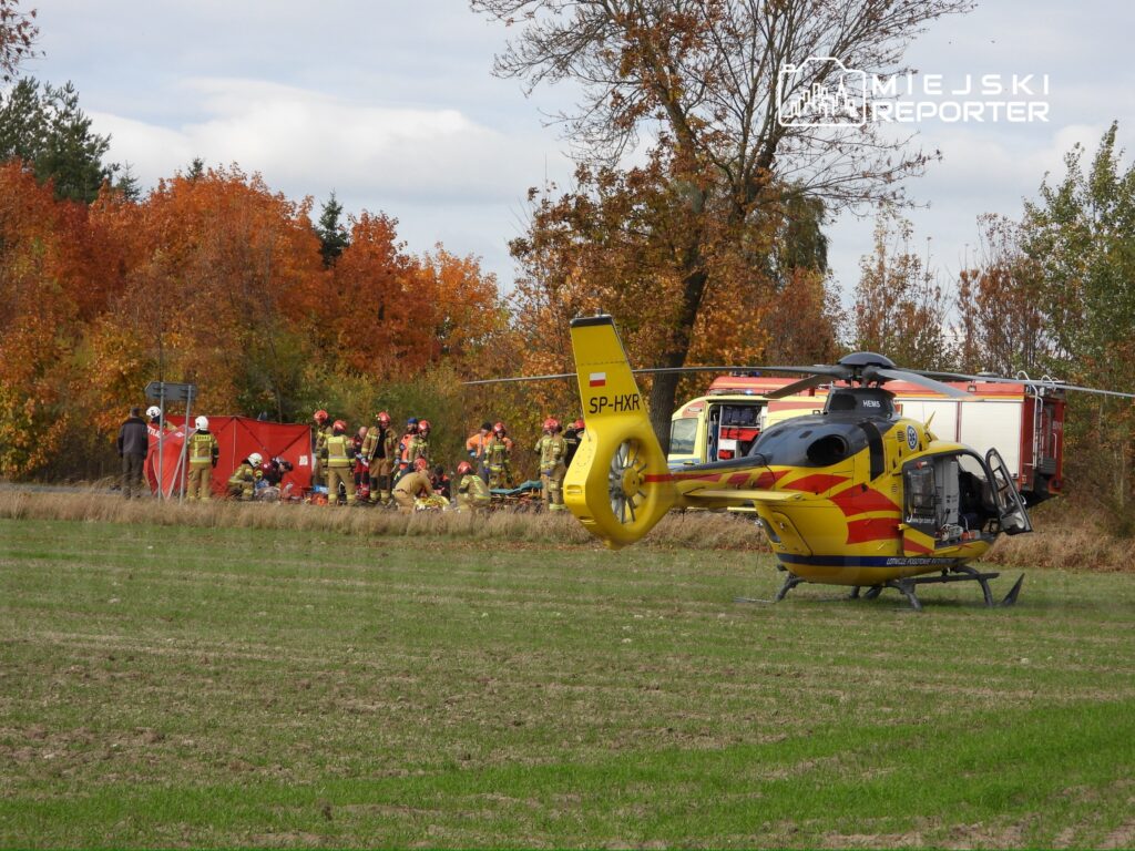 Helikopter ratunkowy lądował na polu obok grupy strażaków pracujących przy akcji ratunkowej. W tle widoczne kolorowe drzewa jesienne.