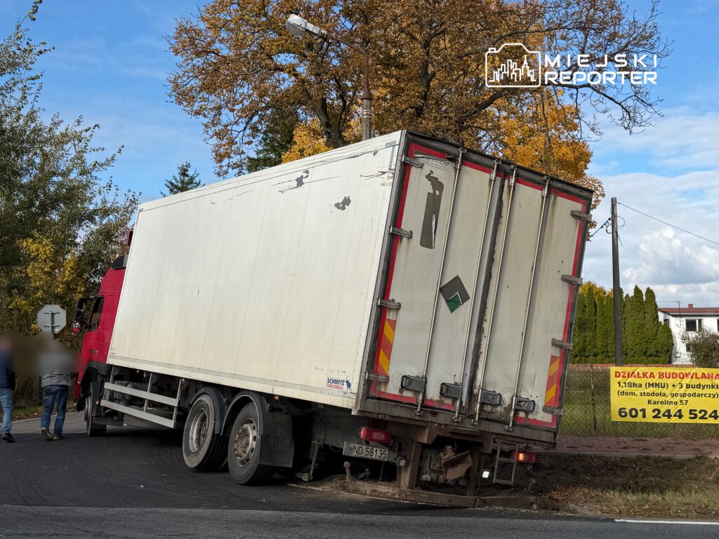 Czerwony ciężarówka z białą naczepą stoi na poboczu drogi, a mężczyźni rozmawiają obok niej.