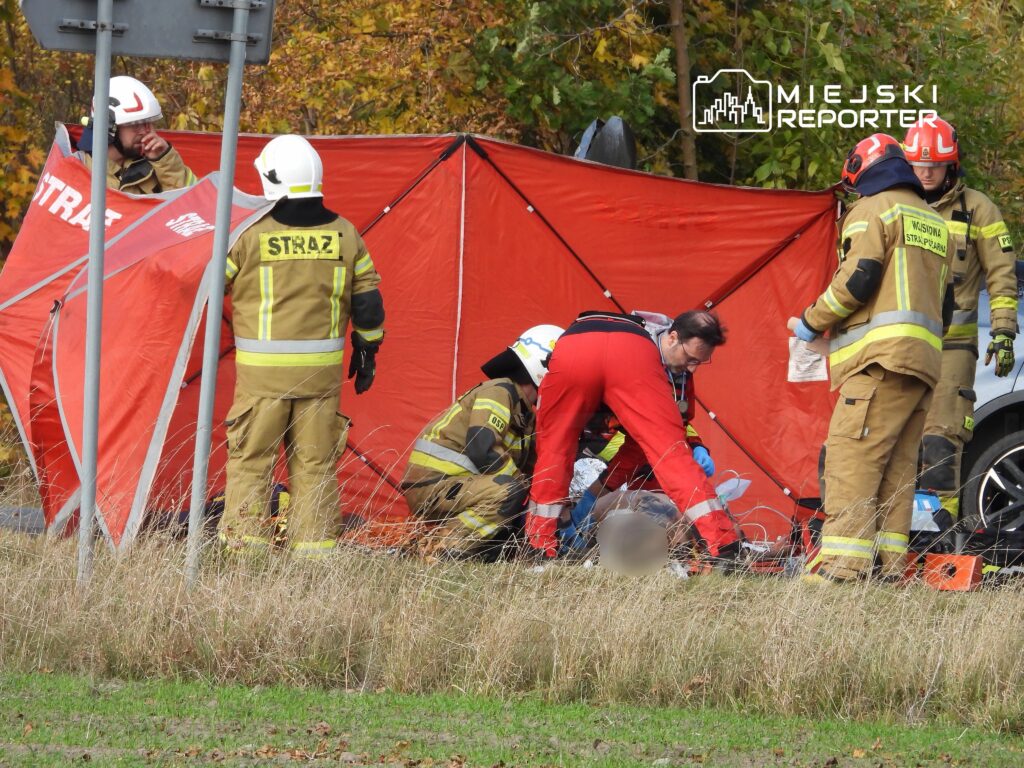 Grupa ratowników w mundurach pracuje nad poszkodowanym w pobliżu czerwonego namiotu na trawiastej łące.