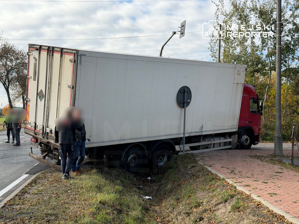 Czterech mężczyzn stoi obok ciężarówki, która wpadła w rów przy drodze, w otoczeniu drzew i znaków drogowych.