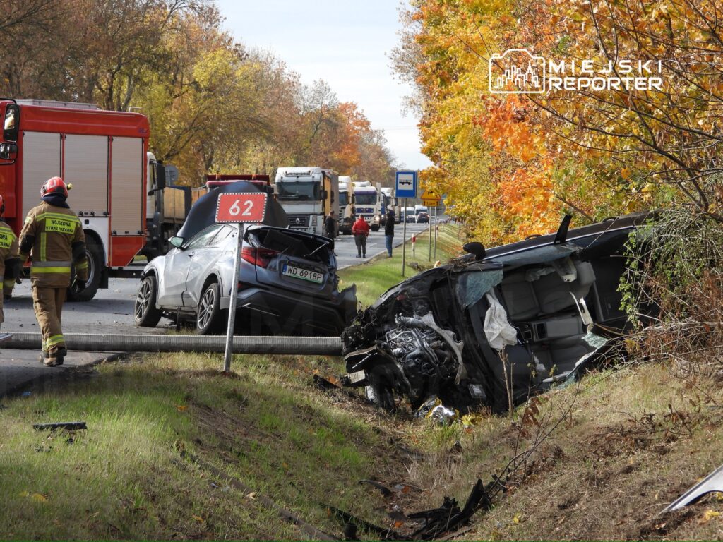Na drodze leży uszkodzony samochód, a obok pracują strażacy w mundurach, w tle widoczne ciężarówki i drzewa w jesiennych kolorach.