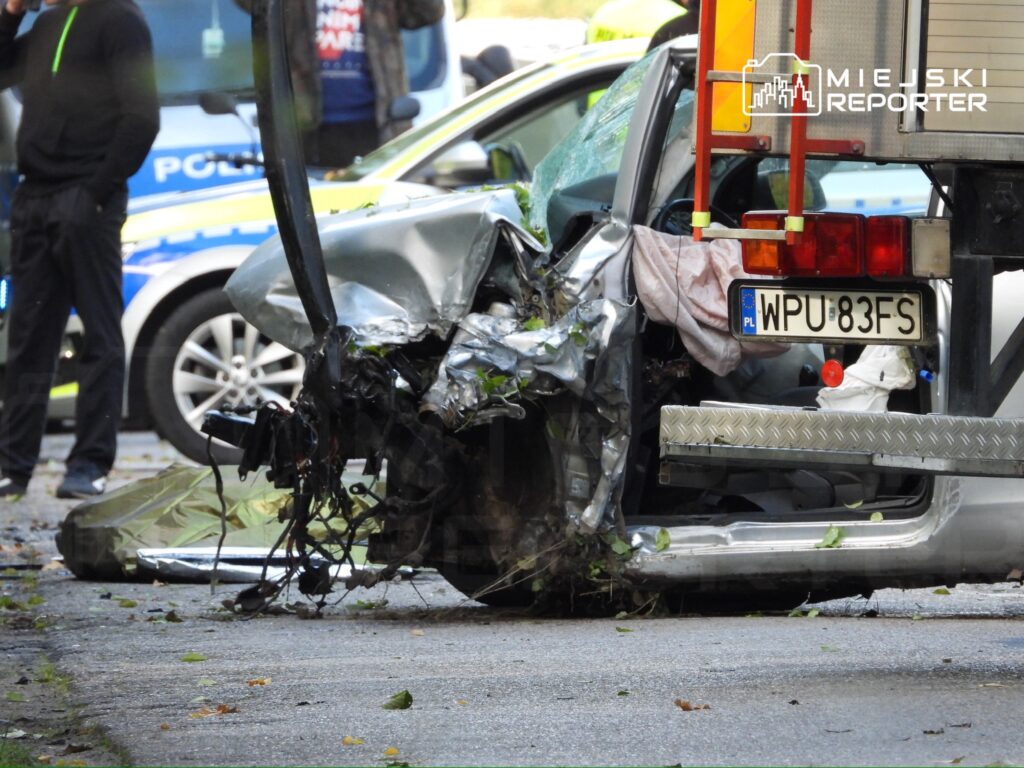 Uszkodzony samochód osobowy z wgniecioną tylną częścią, otoczony przez policjantów i pojazdy służb ratunkowych.