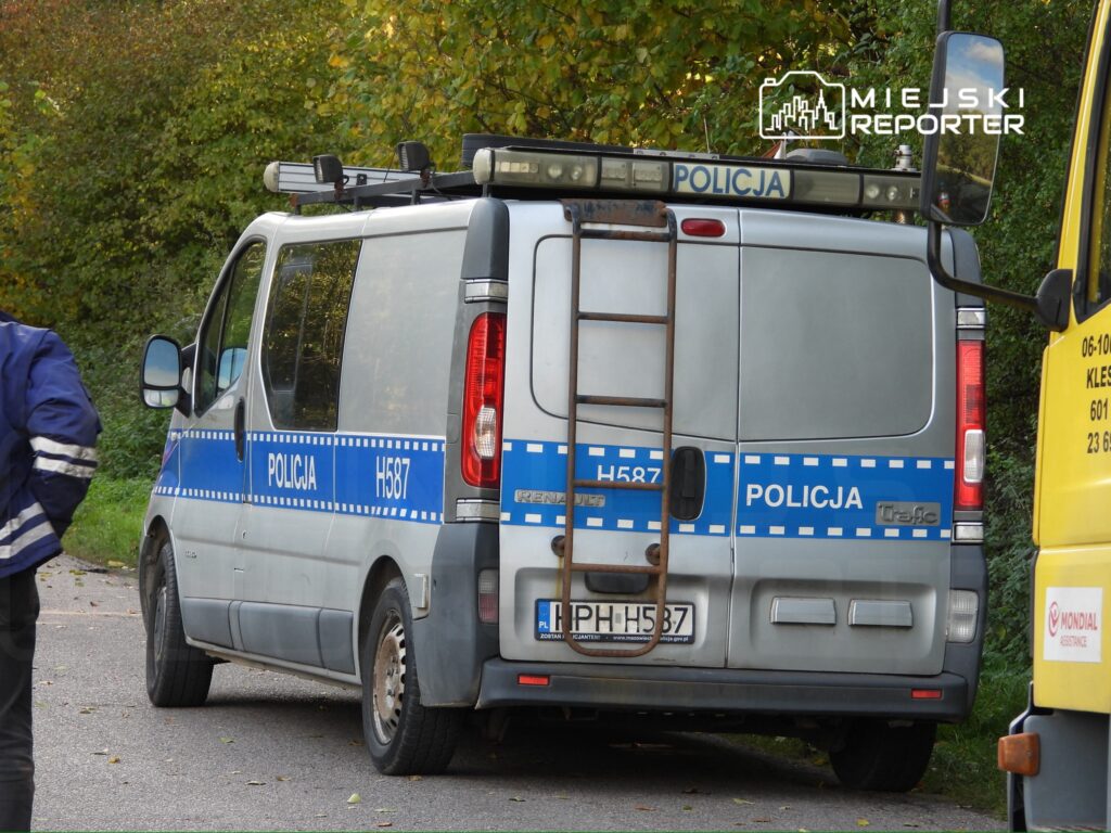 Policyjny samochód z niebieskimi oznaczeniami stoi na poboczu drogi, obok mężczyzny w odblaskowej kurtce.