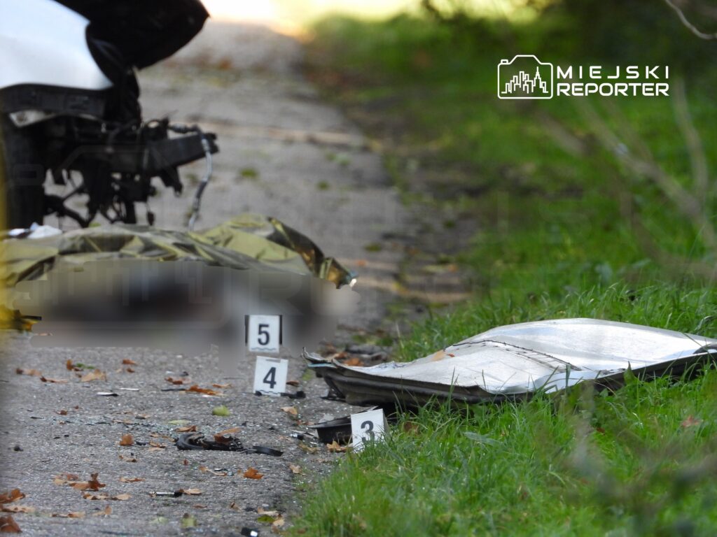 Na drodze leży uszkodzony pojazd, a obok znajdują się oznaczenia i fragmenty rozbitej karoserii.
