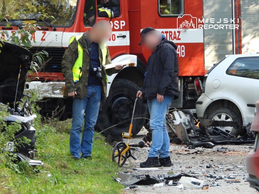 Mężczyzna w kamizelce odblaskowej fotografuje miejsce wypadku, a drugi mężczyzna mierzy odległości przy rozbitych samochodach.