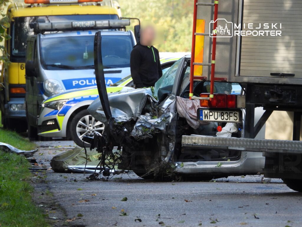 Zniszczony samochód osobowy stoi przy drodze, obok pojazdów policji i straży, w otoczeniu opadłych liści.