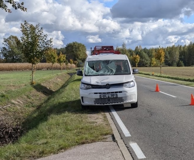 Biały samochód dostawczy z uszkodzoną szybą stoi na poboczu drogi, obok pomarańczowe trójkąty ostrzegawcze.