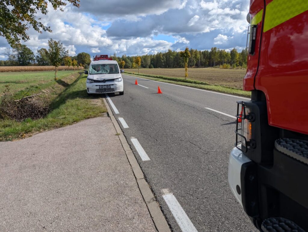 Biały samochód osobowy zatrzymany na poboczu drogi, obok pomarańczowe pachołki, w tle widok na pola i las.