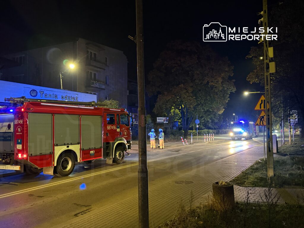 Czerwony wóz strażacki stoi na ulicy, obok grupy pracowników w odblaskowych kamizelkach, w nocy.