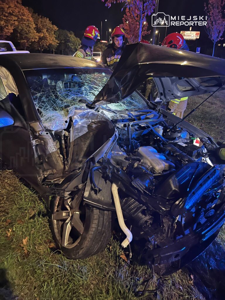 Uszkodzony samochód osobowy z rozbitą szybą i wgniecioną karoserią stoi na poboczu, obok strażacy w hełmach.