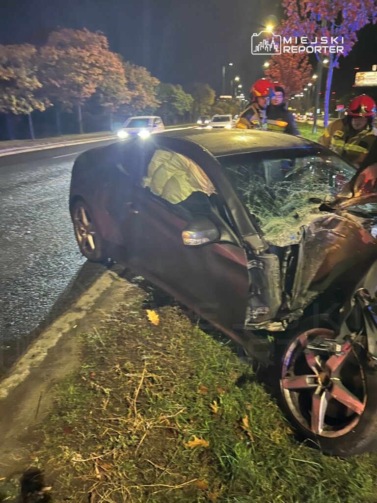 Czarny samochód osobowy z uszkodzoną karoserią i włączonymi światłami awaryjnymi stoi na poboczu drogi, obok pracują strażacy.