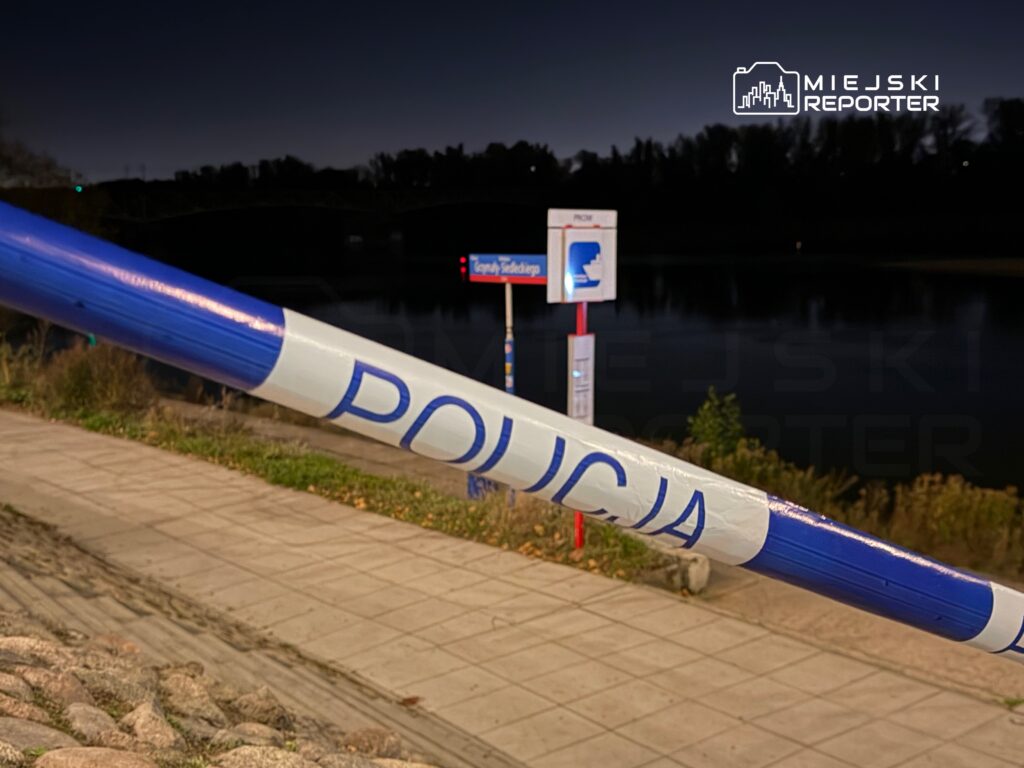 Policja zabezpiecza teren przy rzece, na tle słupa z informacją o przystanku. Nocna sceneria.