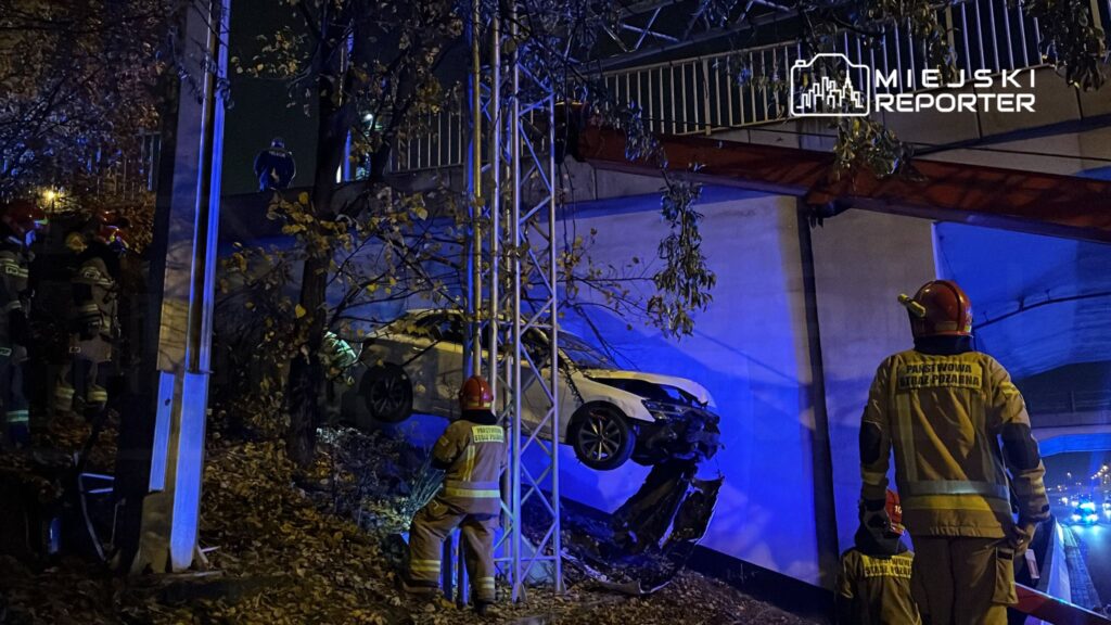 Grupa strażaków interweniuje przy wypadku drogowym, gdzie samochód zawisł nad skarpą, otoczony niebieskimi światłami.