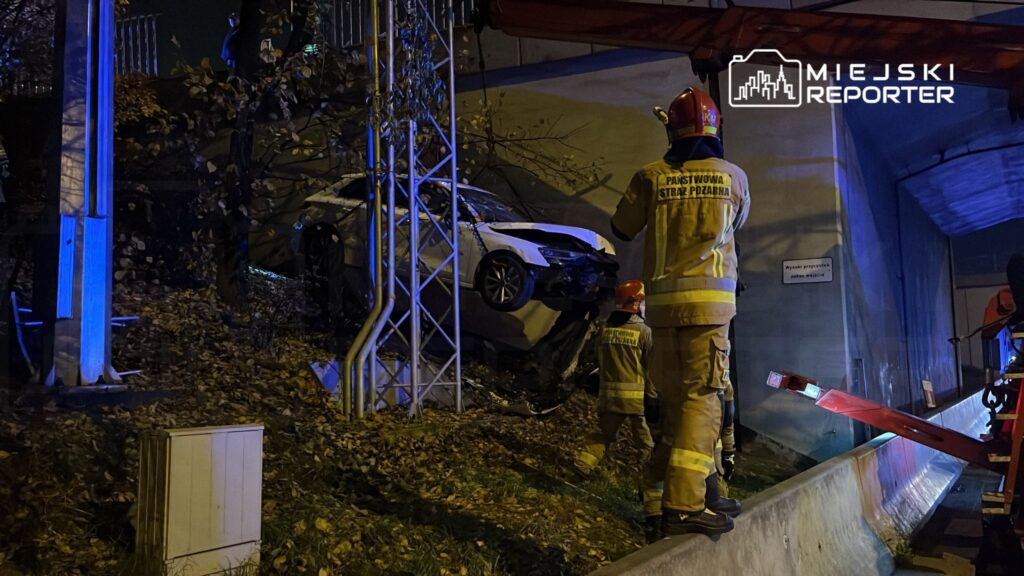 Strażacy w mundurach z logo Państwowej Straży Pożarnej zabezpieczają wrak samochodu zawieszonego na skarpie.