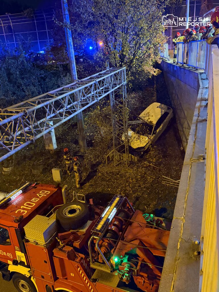 Strażacy przy wozie strażackim, interweniujący przy wypadku samochodowym w wąwozie, otoczonym drzewami i oświetleniem.