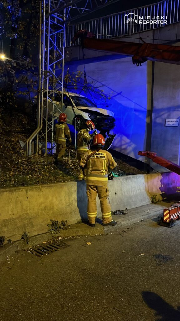 Grupa strażaków w hełmach pracuje nad usunięciem samochodu z wąskiego zbocza w nocy, oświetlona niebieskimi światłami.