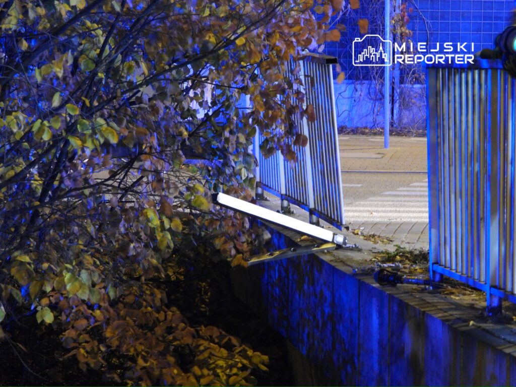 Oświetlona lampa LED umieszczona na krawędzi muru w miejskim otoczeniu, otoczona jesiennymi liśćmi.