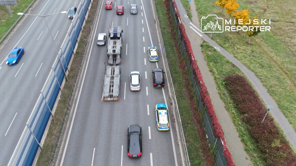 Policyjny radiowóz zatrzymany na poboczu ruchliwej drogi, obok ciężarówki i osobowych samochodów.