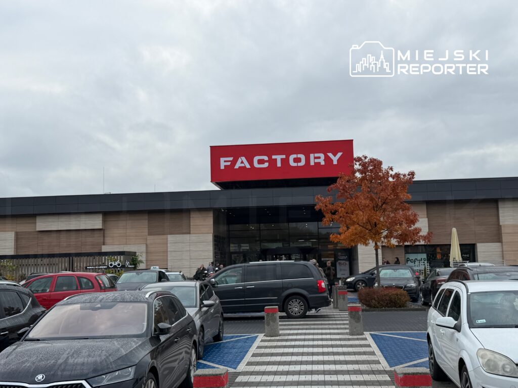 Centrum handlowe z dużym napisem "FACTORY" na fasadzie, otoczone zaparkowanymi samochodami i drzewem w jesiennych kolorach.