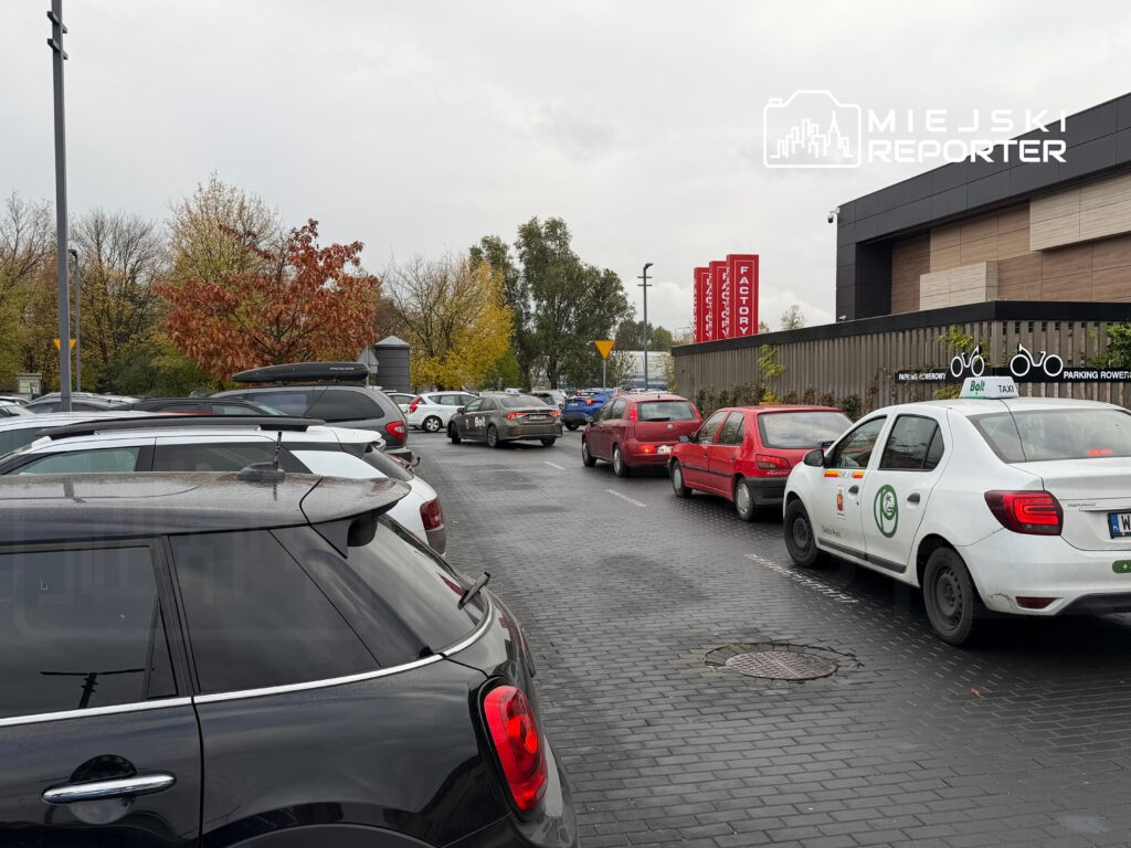 Parking z dużą ilością zaparkowanych samochodów, w tym taksówki i czerwone auta, otoczony drzewami w jesiennej scenerii.