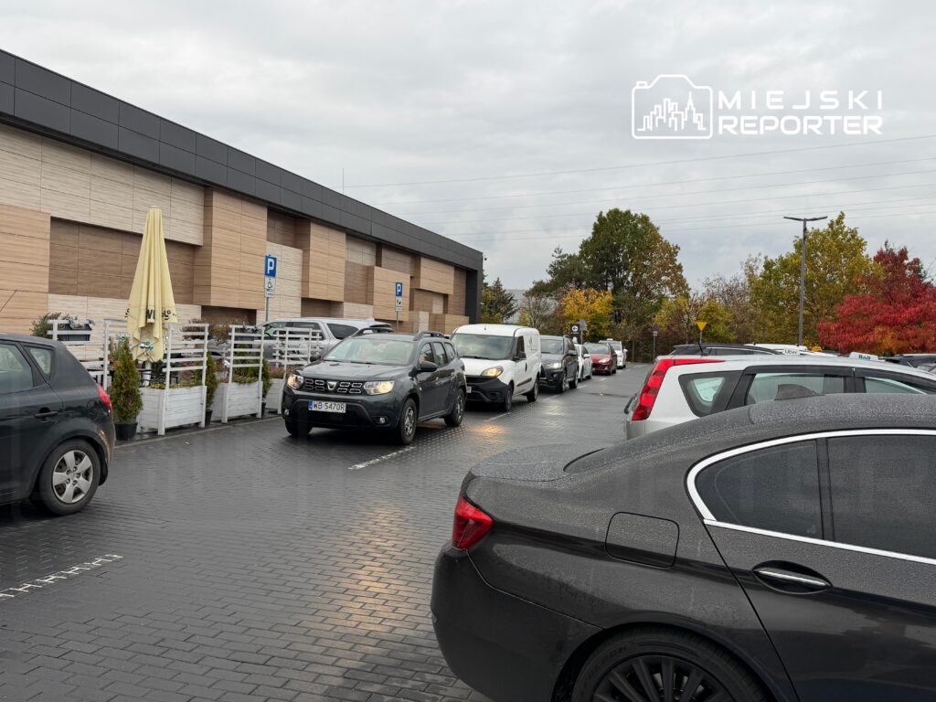 Ruch samochodowy na parkingu przed nowoczesnym budynkiem, z widocznymi zaparkowanymi pojazdami i drzewami w tle.