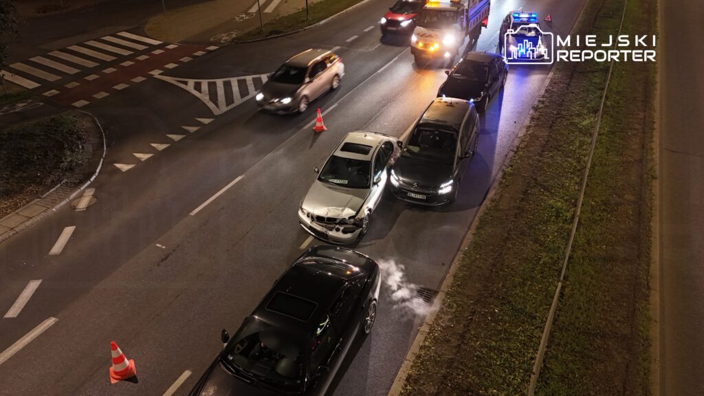 Wielu kierowców zatrzymanych w korku na drodze po kolizji dwóch samochodów w nocy. Widać radiowóz i pojazd z włączonymi światłami.