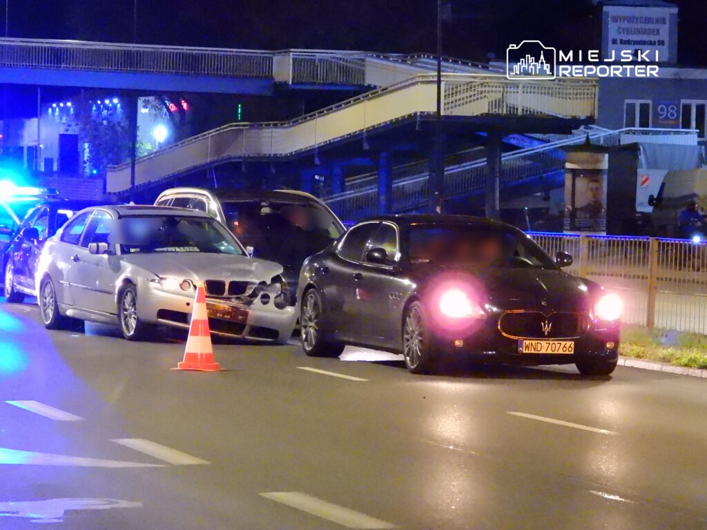Zderzenie dwóch samochodów na nocnej ulicy, z uszkodzonym pojazdem w kolorze srebrnym i czarnym samochodem sportowym.