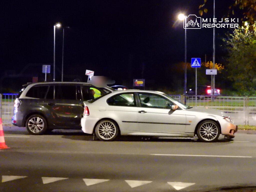 Dwa samochody, jeden szary SUV i jeden biały sedan, stoją obok siebie na drodze nocą, przy włączonych światłach.