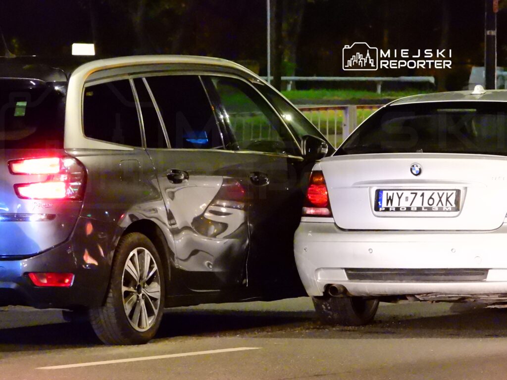 Dwa samochody osobowe, czarny SUV i biały sedan, zderzyły się na drodze nocą. Widać uszkodzenia na bocznej części SUV-a.