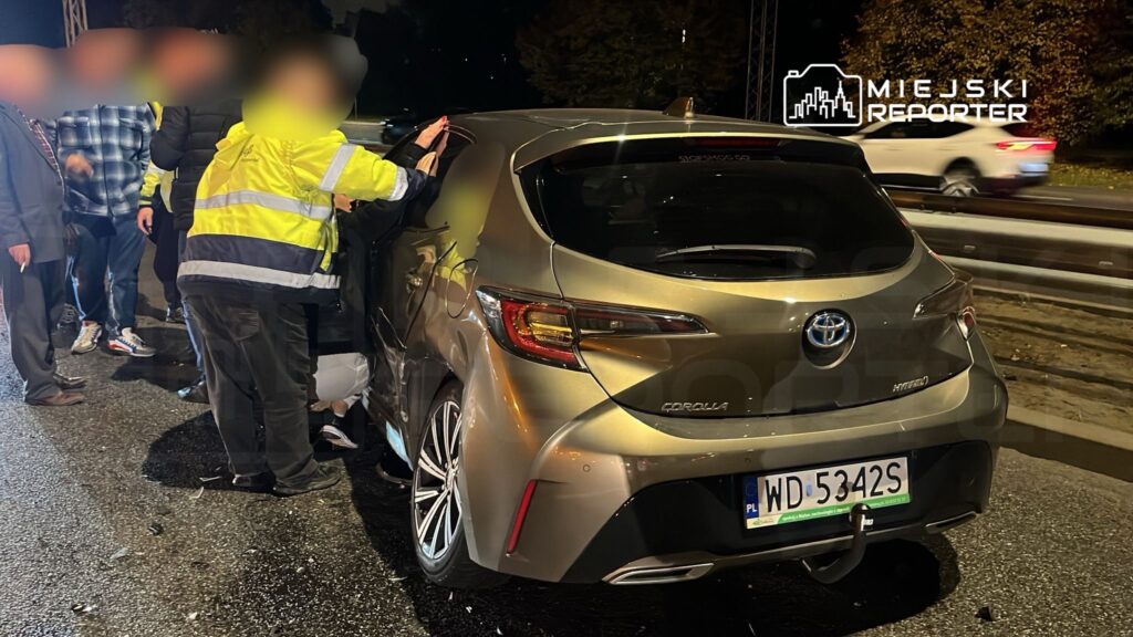 Grupa osób w odblaskowych kamizelkach sprawdza uszkodzenia samochodu osobowego na drodze w nocy.