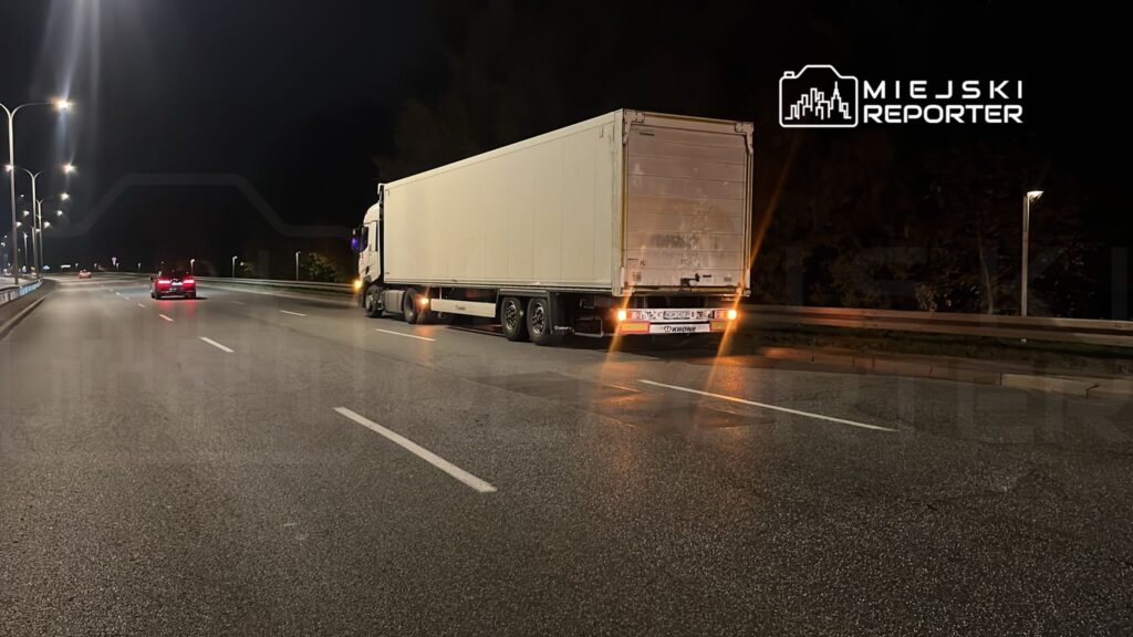 Czarny samochód osobowy porusza się obok białej ciężarówki stojącej na oświetlonej drodze nocą.