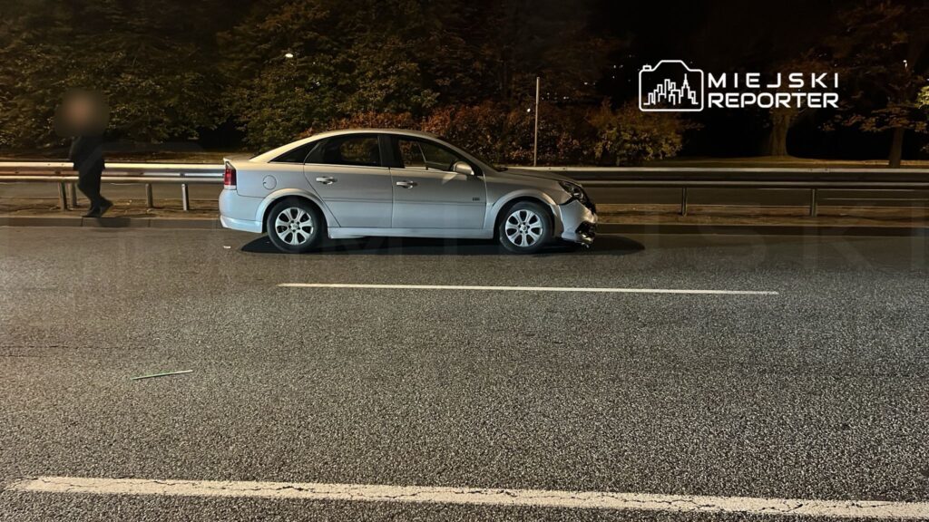 Na jezdni stoi uszkodzony srebrny samochód osobowy, obok idzie pieszy w ciemnym ubraniu.