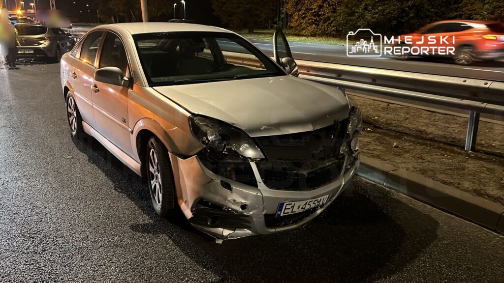 Uszkodzony srebrny samochód osobowy stoi na poboczu drogi, z otwartymi drzwiami i widocznymi uszkodzeniami przodu.