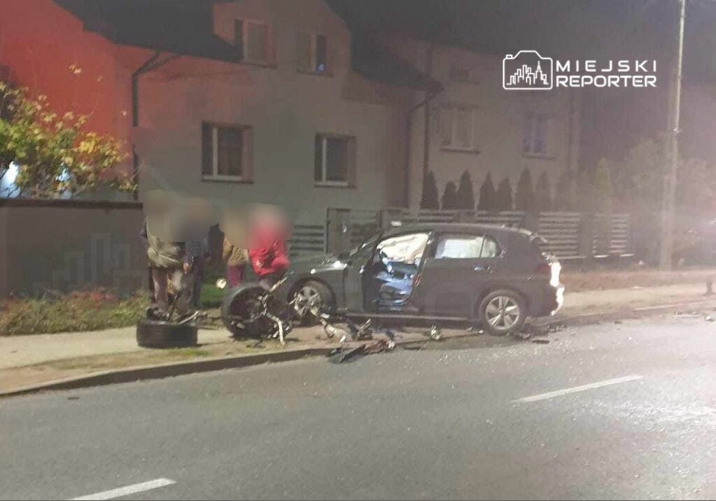 W nocy, w pobliżu uszkodzonego samochodu, grupa ludzi bada skutki wypadku drogowego. Na jezdni leżą części roweru.