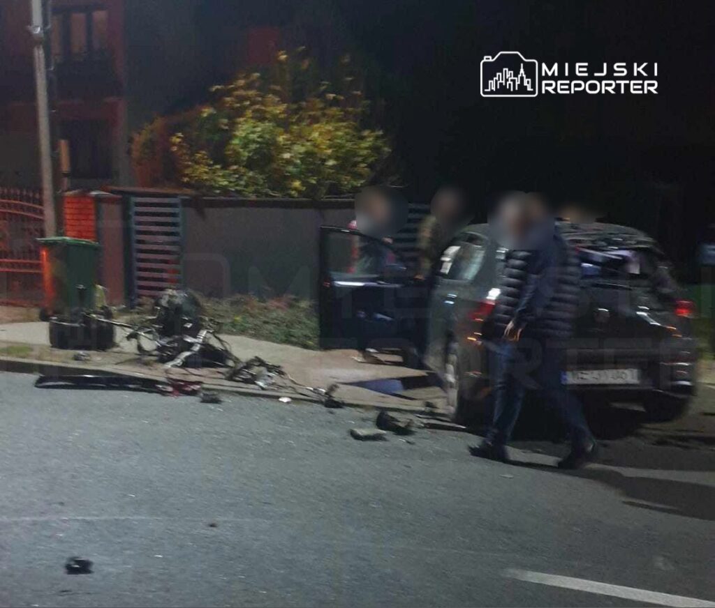 Grupa osób przy uszkodzonym samochodzie na ulicy, obok leżące fragmenty pojazdu i narzędzia.