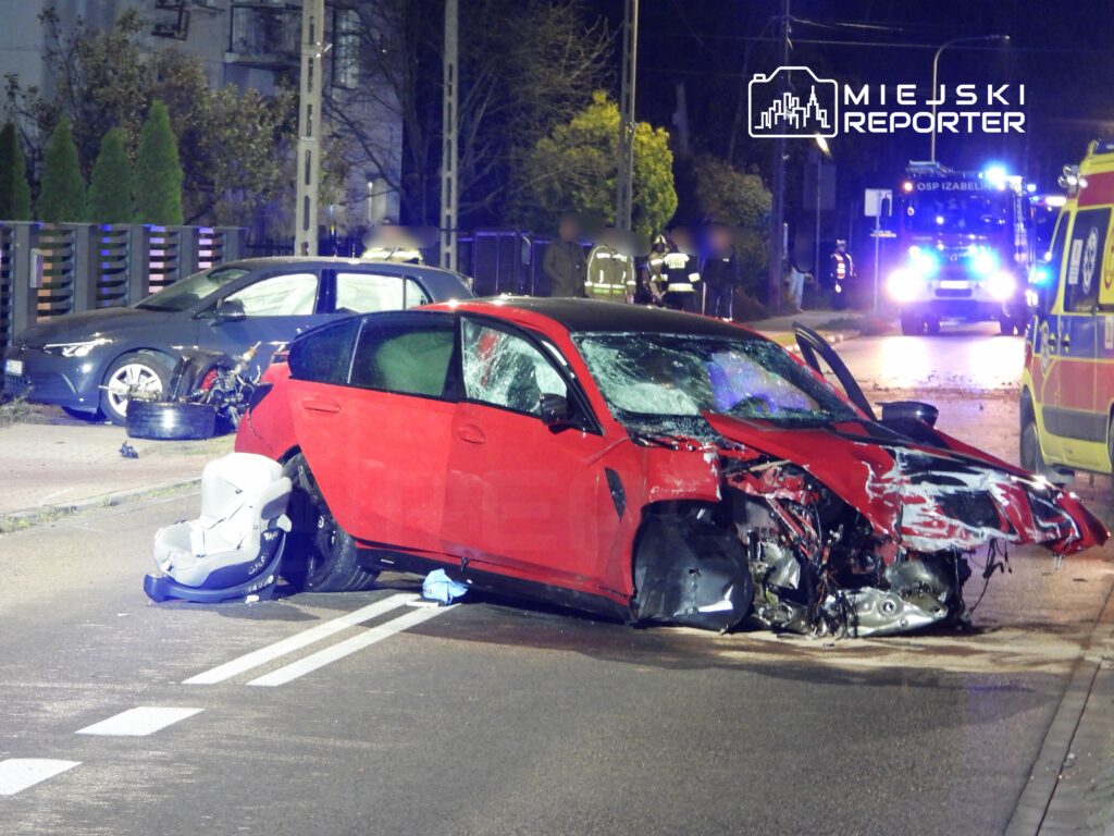 Czerwony samochód osobowy z uszkodzoną karoserią stoi na drodze, obok widać wózek dziecięcy oraz pojazdy służb ratunkowych.