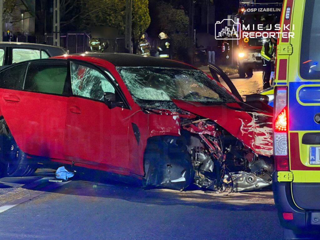 Czerwony samochód osobowy z poważnymi uszkodzeniami po wypadku drogowym na ulicy, obok stoją pojazdy ratunkowe.