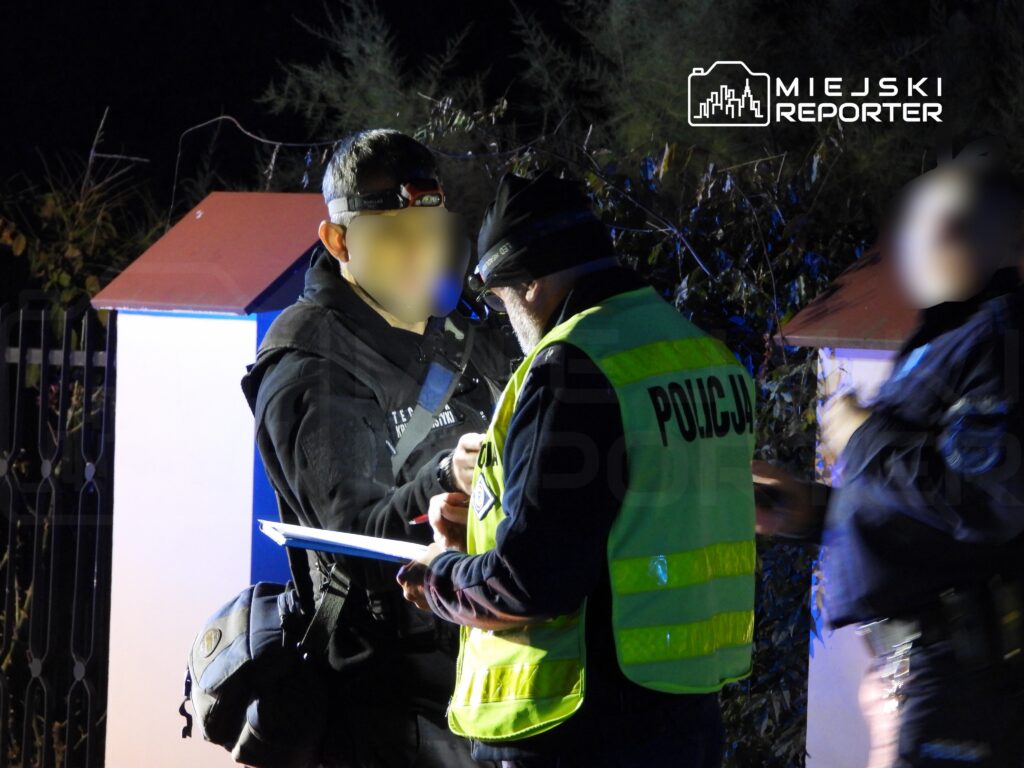 Mężczyzna w ciemnym ubraniu rozmawia z policjantem w żółtej kamizelce odblaskowej w nocy przy ogrodzeniu.