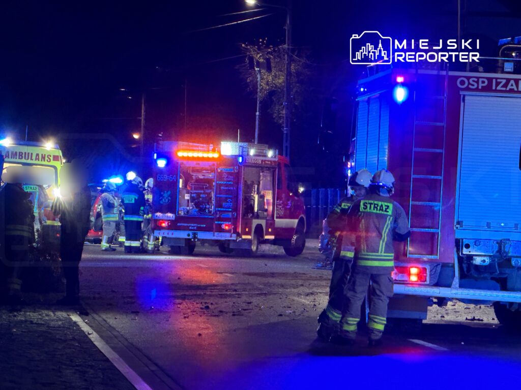 Grupa strażaków w mundurach omawia sytuację przy wozie strażackim, obok ambulansu na oświetlonej ulicy nocą.