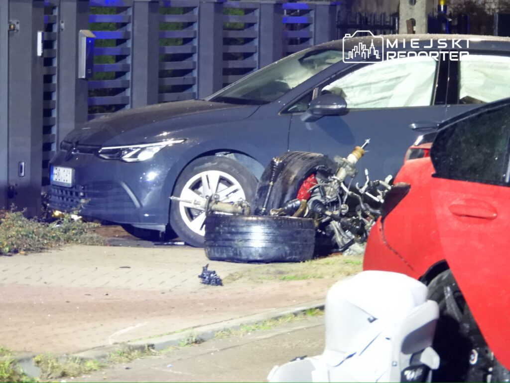 Zdemolowany motocykl leży na chodniku obok zaparkowanego samochodu osobowego w nocy.
