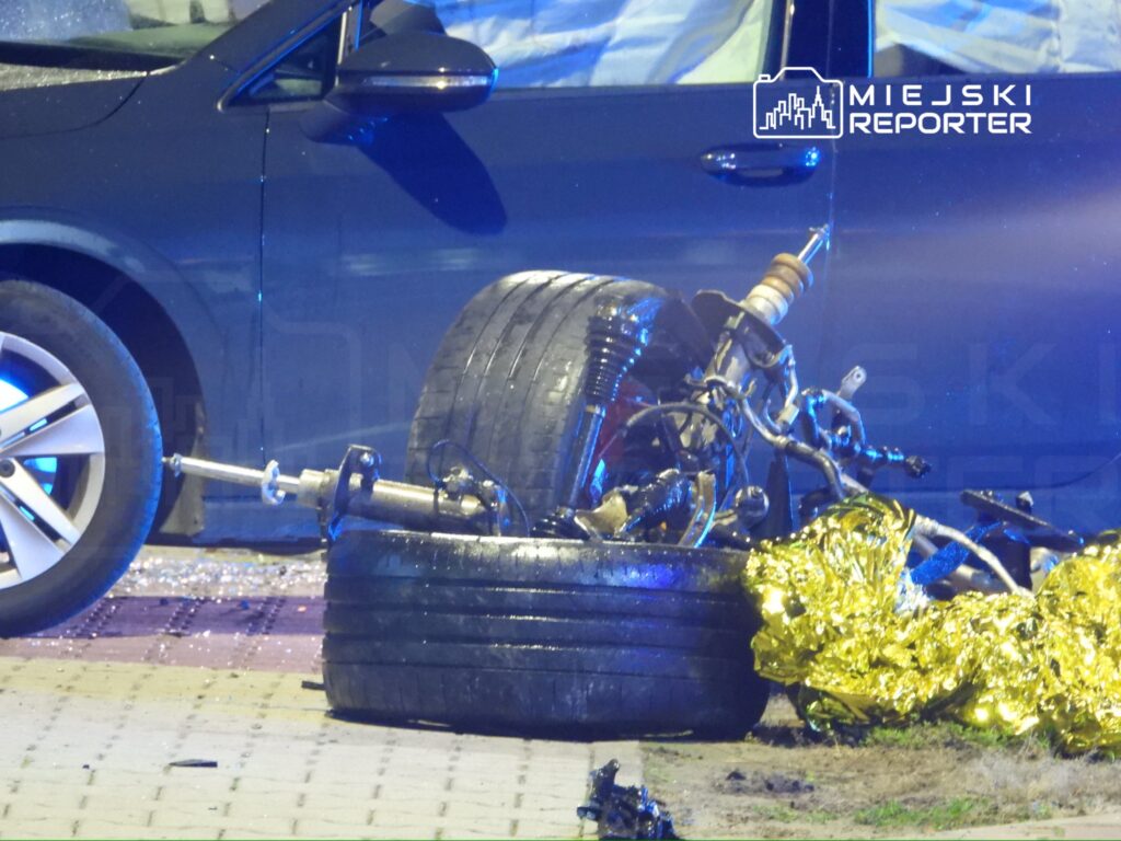 Zniszczony motocykl leży na jezdni obok ciemnego samochodu, otoczony przez policyjne światła i fragmenty szkła.