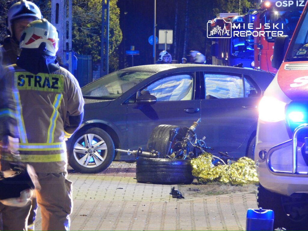 Strażacy w mundurach rozmawiają obok uszkodzonego samochodu na jezdni, obok leży motocykl z odłączonymi kołami.