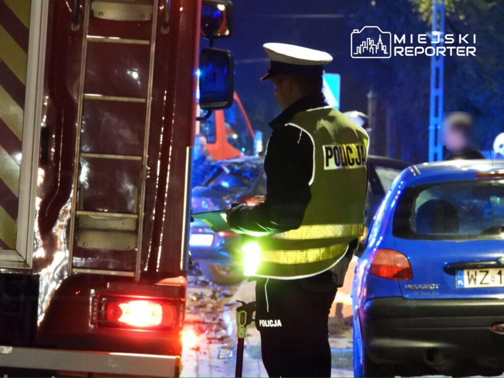 Policjant w odblaskowej kamizelce kontroluje miejsce wypadku drogowego w nocy, obok wozu strażackiego.