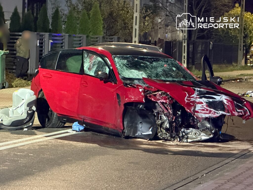 Czerwony samochód osobowy z uszkodzoną przednią częścią stoi na jezdni, obok fotelik dziecięcy.