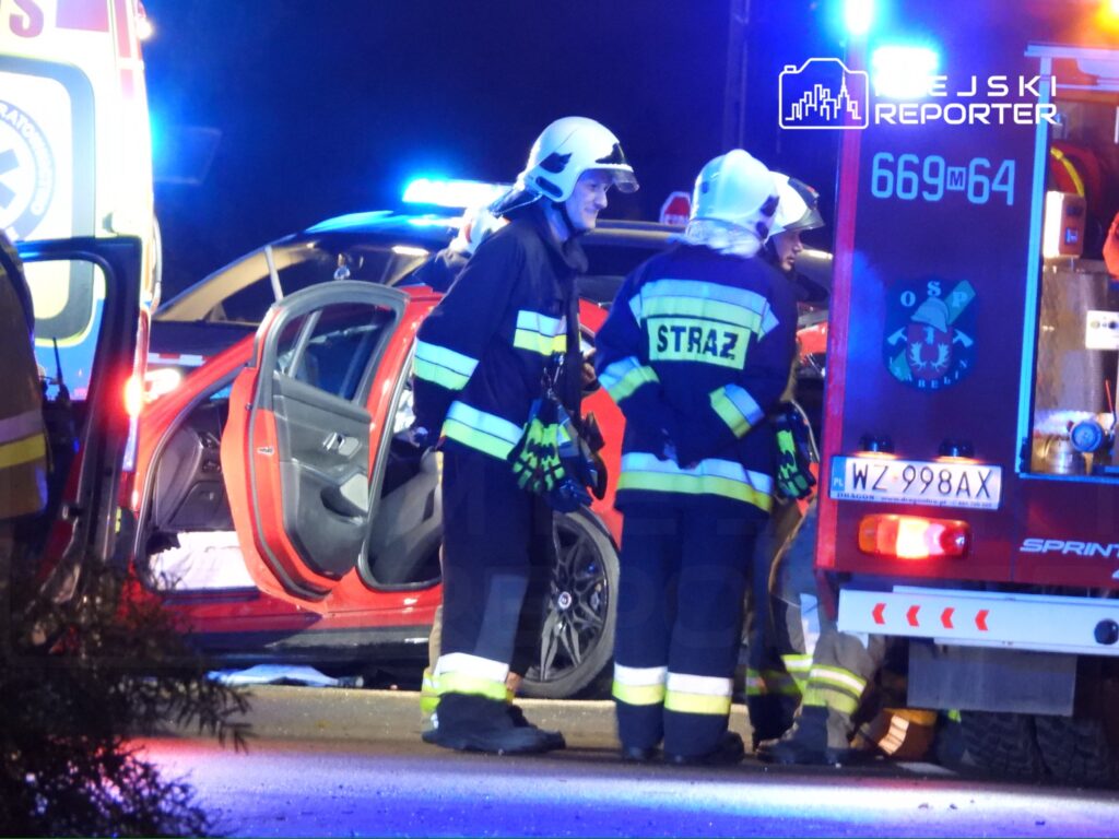 Strażacy w mundurach rozmawiają obok uszkodzonego czerwonego samochodu, w tle widać karetkę z włączonymi sygnalizatorami.